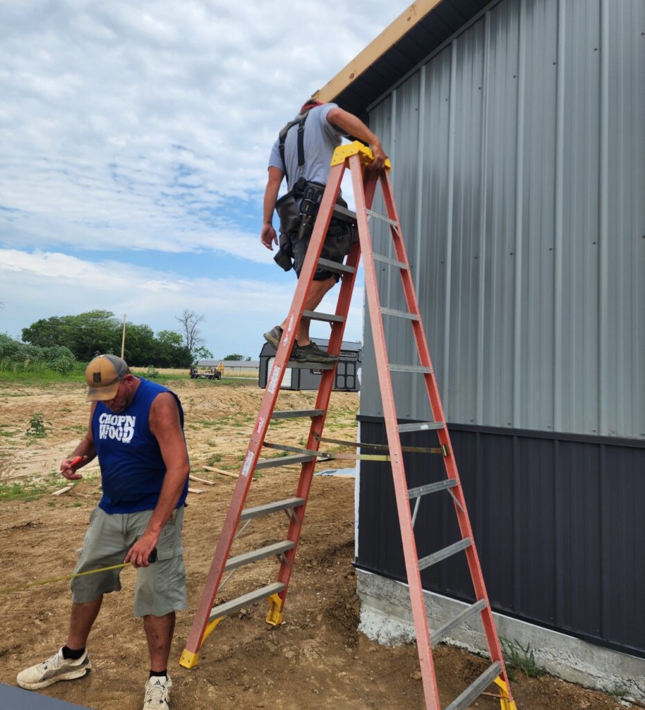 An action shot of two construction workers at a work site, focusing on the exterior installation of a metal building.