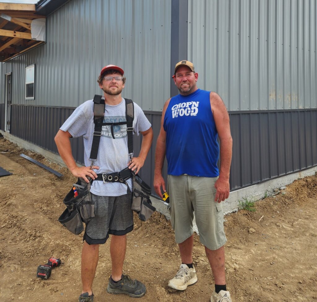 A candid outdoor photograph featuring two male construction workers standing in front of a newly installed grey metal building exterior.