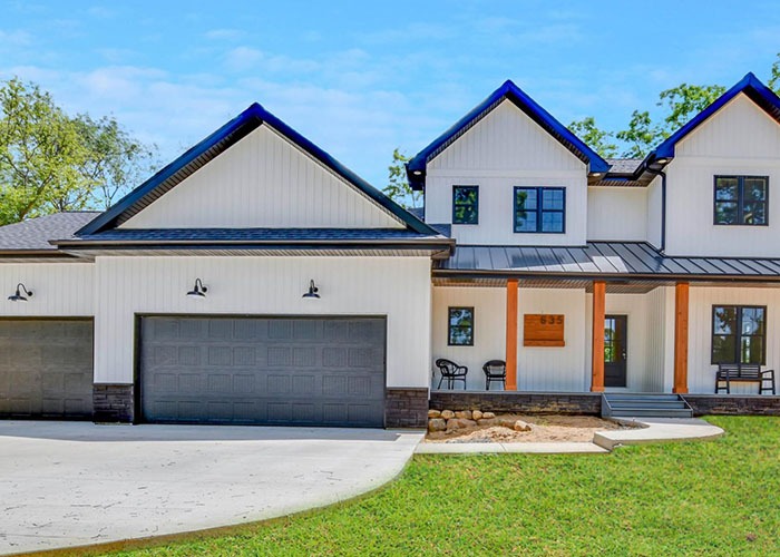 Modern white farmhouse with black trim by DreamStart Building Company, featuring a three-car garage, natural wood porch pillars, and a sleek metal roof design.